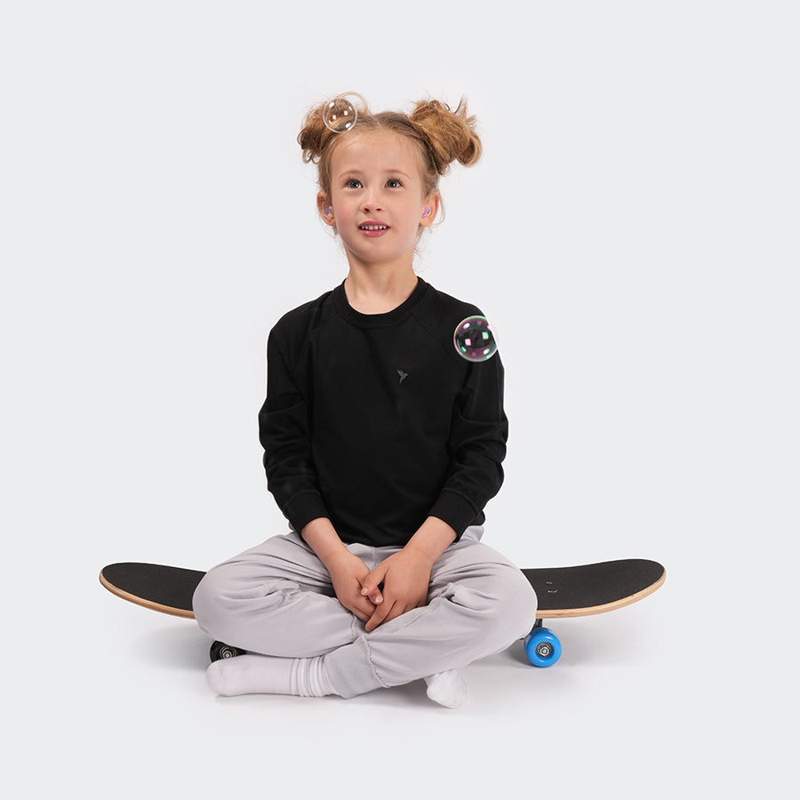 Model sat on skateboard with bubbles floating, wearing Black Rare Kids Long Sleeve Tshirt & grey joggers - sensory friendly & made for the neurodivergent community on grey background